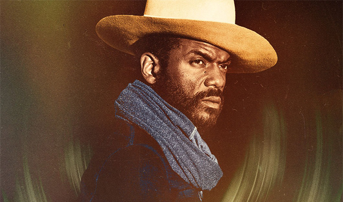 A man wearing a tan cowboy hat and blue scarf looks intensely to the side against a moody, dark green background.