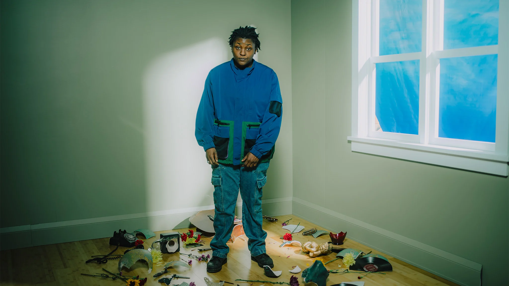 Person in a blue jacket and jeans stands in a corner room surrounded by scattered flowers, vinyl records, and misc objects.