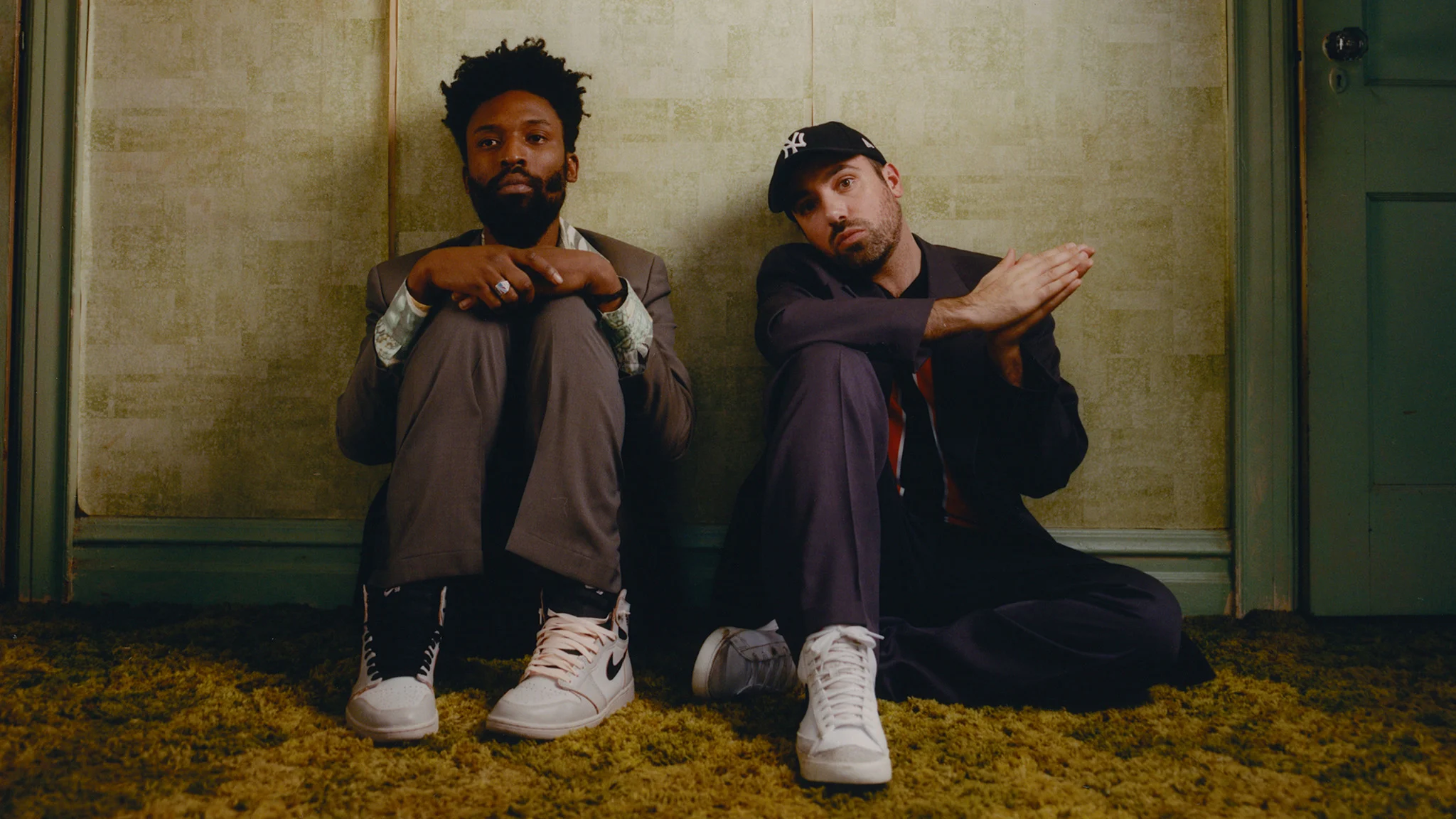 Two men sit on a green carpet against a wall, wearing suits and white sneakers, one in a NY cap, looking at the camera.