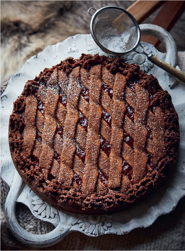 Chocolate tart with lattice top dusted with sugar on a decorative white platter, with a small sifter nearby.