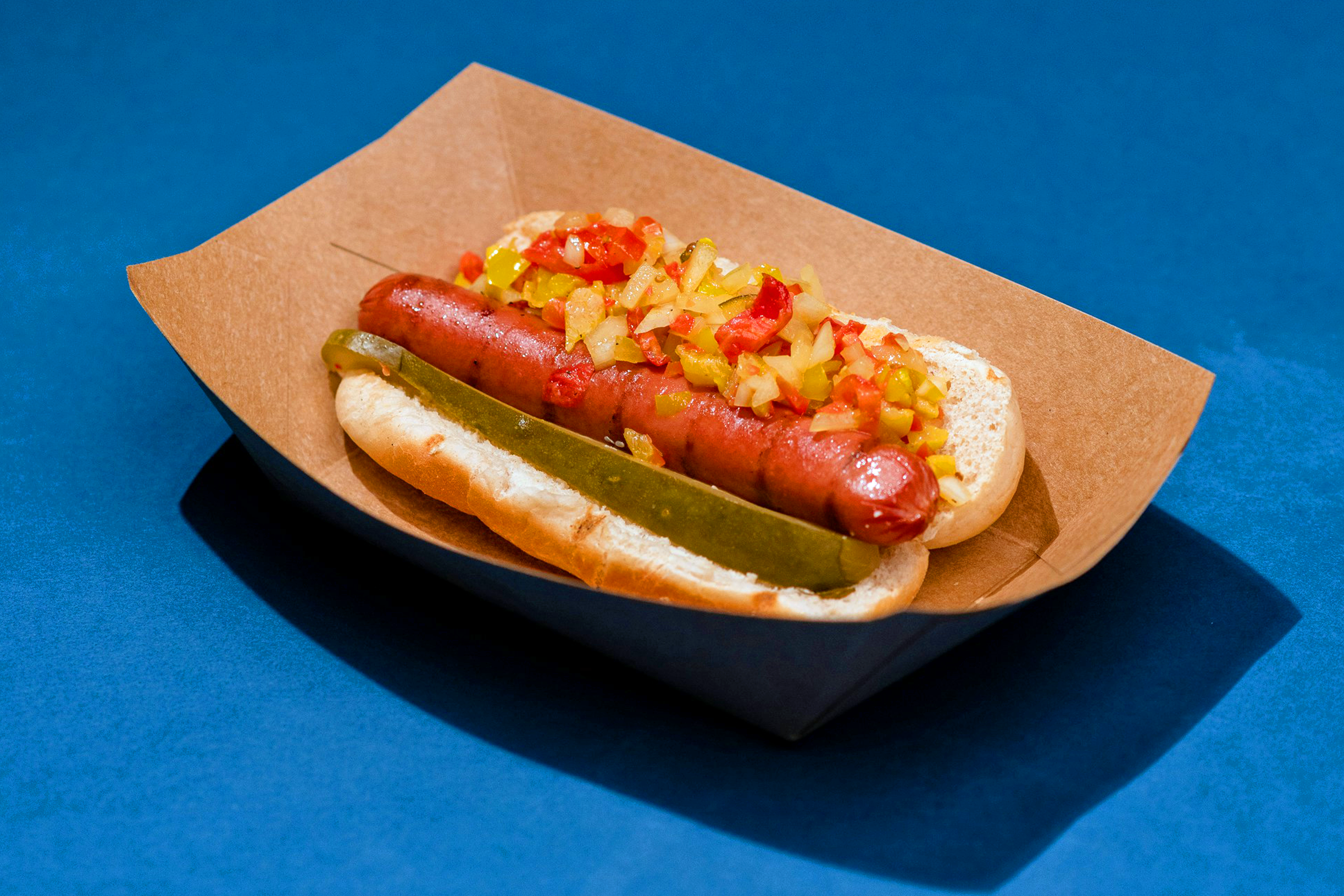 A hot dog in a bun topped with a pickle, diced onions, and red and yellow peppers, served in a kraft paper tray.