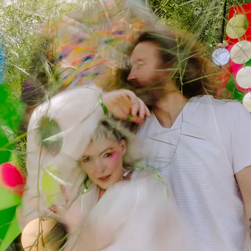 Two people lying in colorful greenery, surrounded by vibrant decorations, captured with a dreamy, motion-blurred effect.