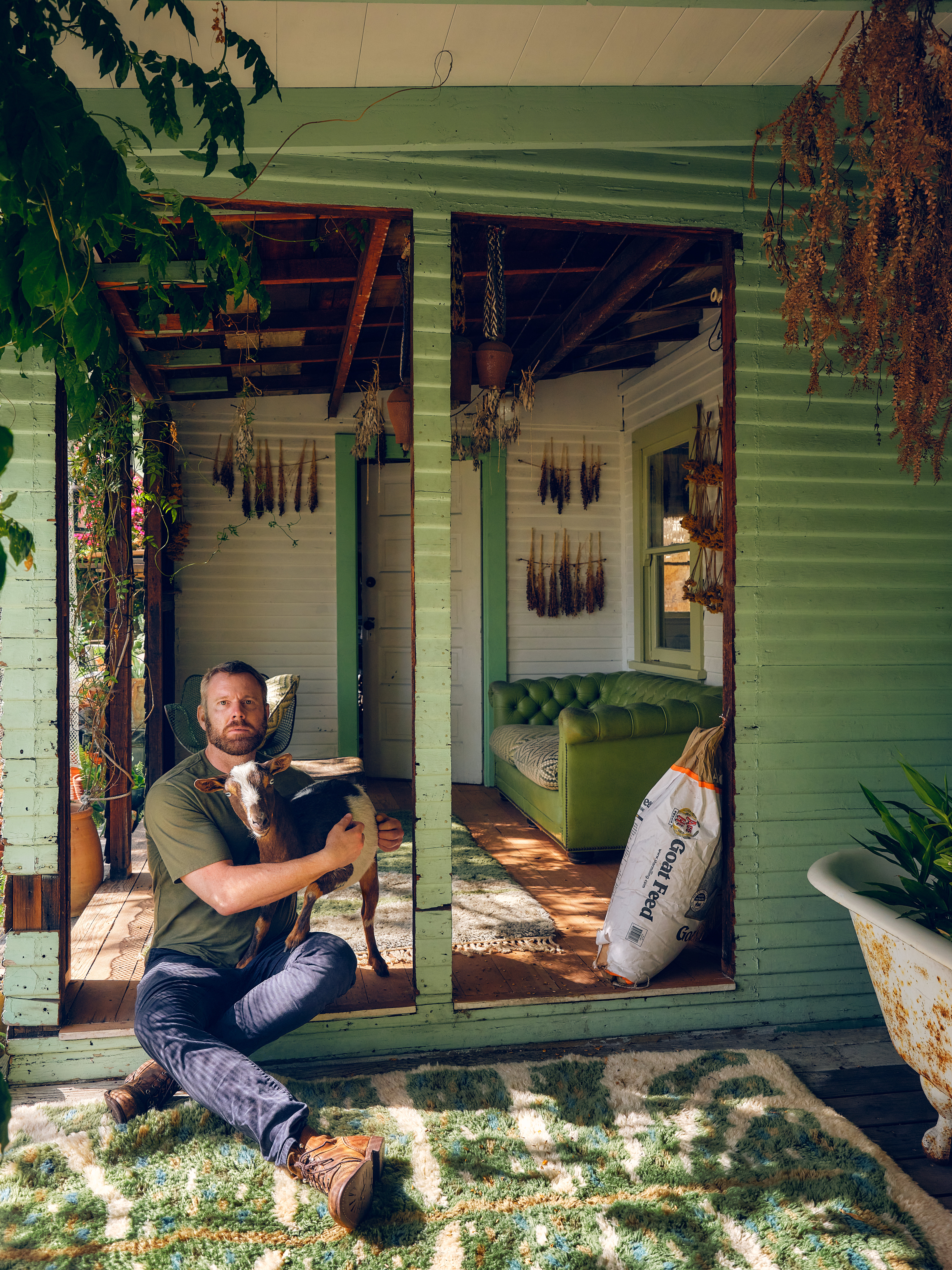 A man sits on a green porch holding a small goat, with dried herbs hanging and a bag of goat feed nearby.