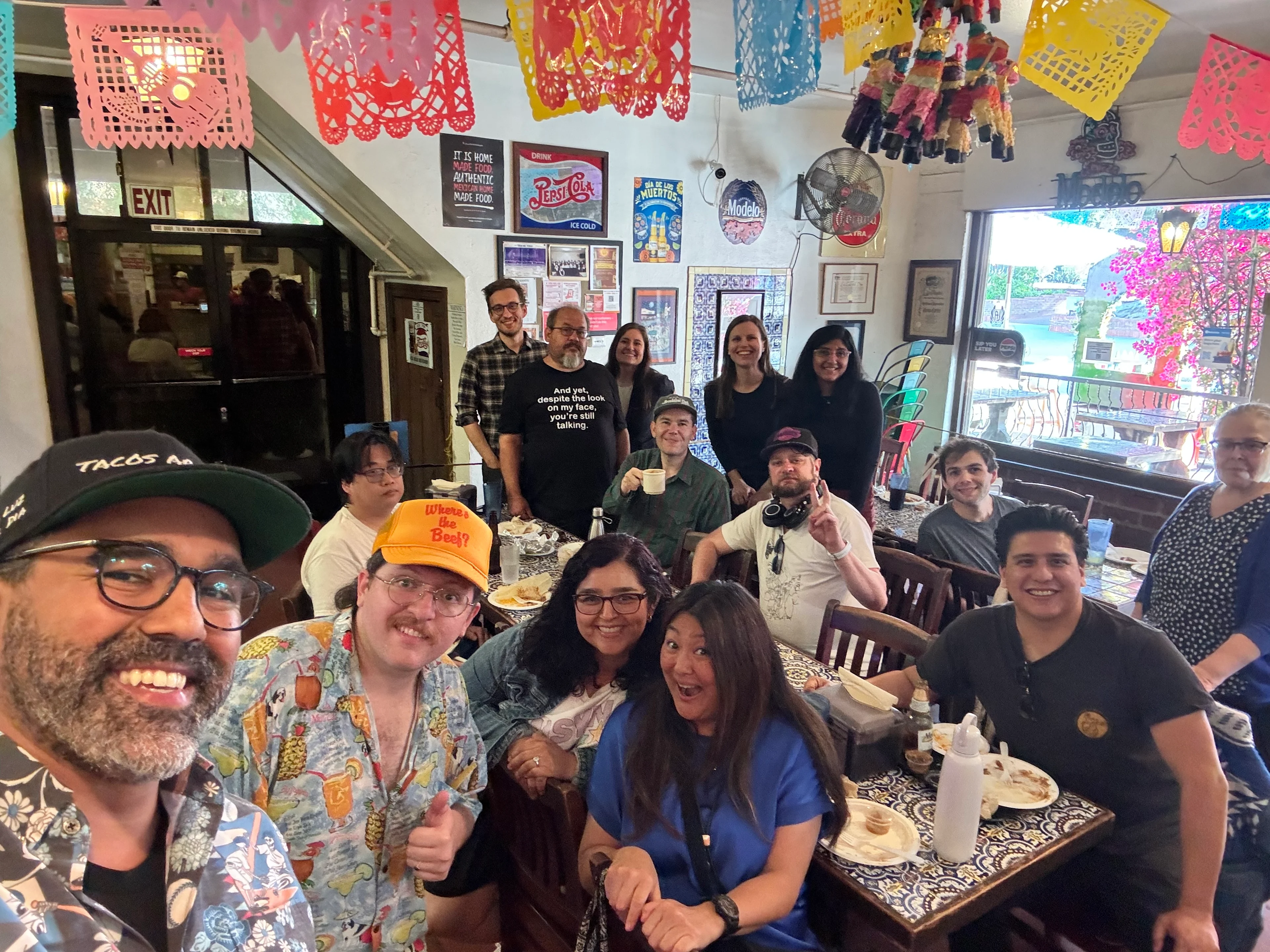 A man takes a selfie in a colorful restaurant surrounded by friends and family enjoying the food. 