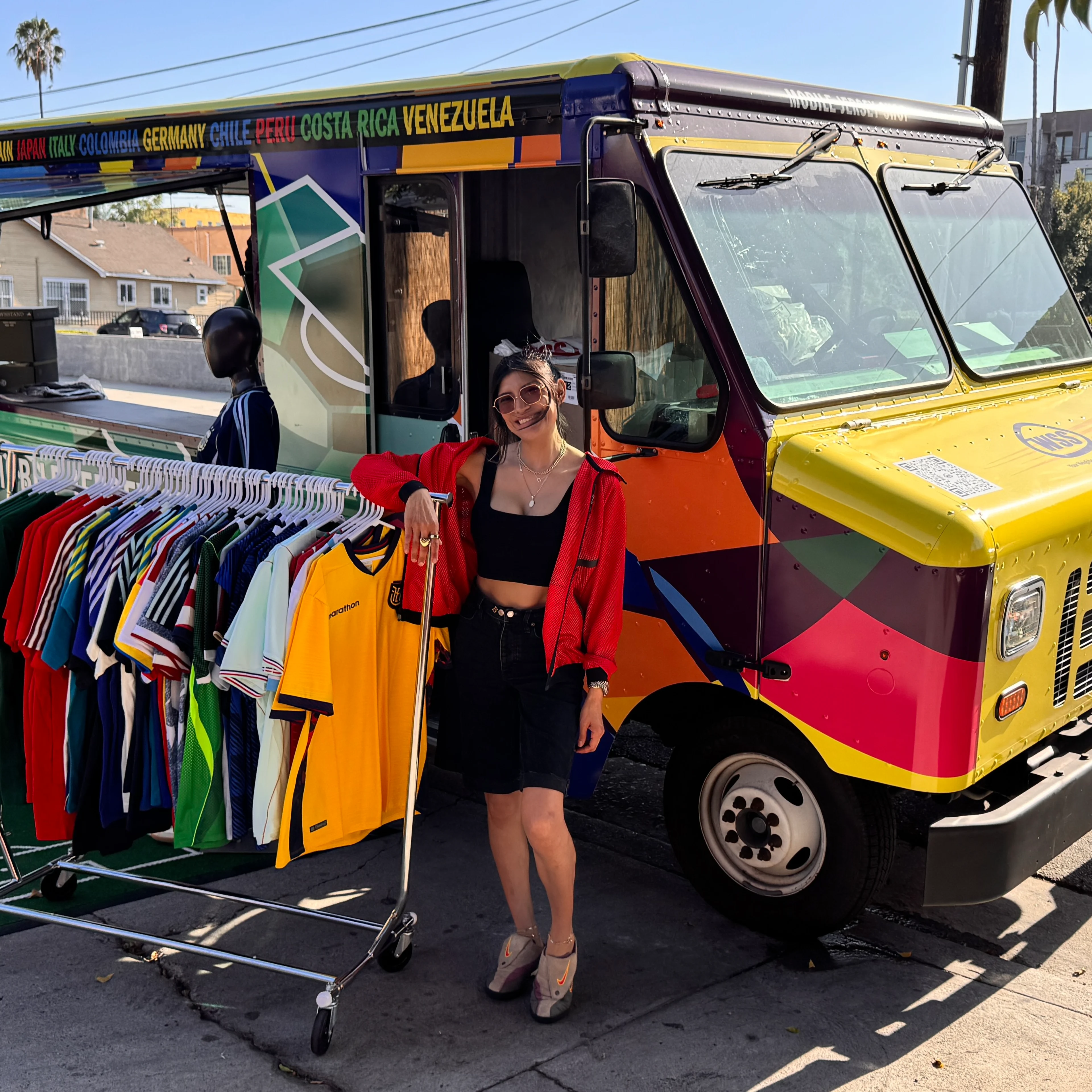 A smiling woman in a red jacket stands beside a colorful mobile jersey truck with a rack of soccer shirts on a sunny street.