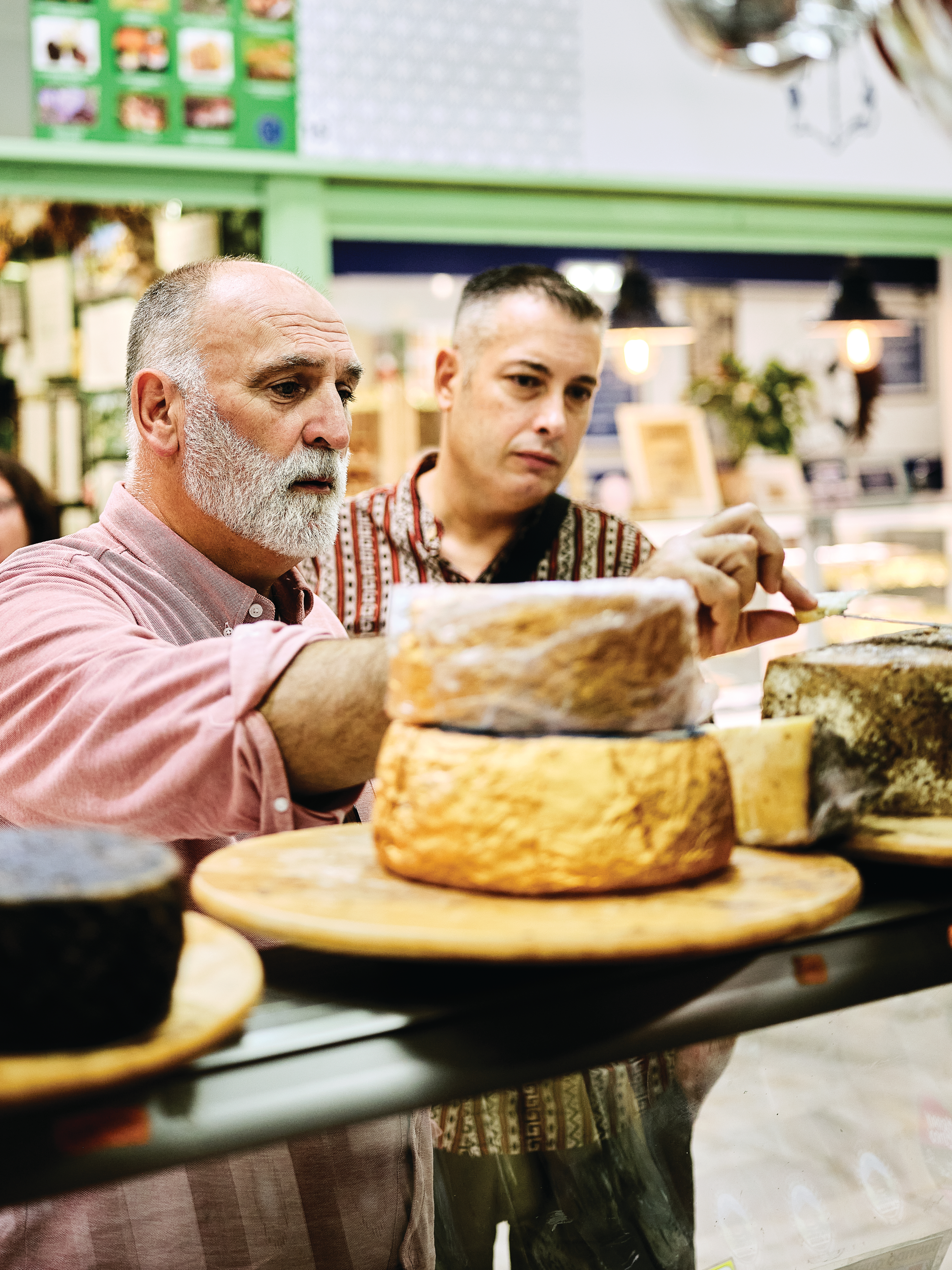 Two men examine large wheels of aged cheese on a wooden board at an indoor market cheese counter.