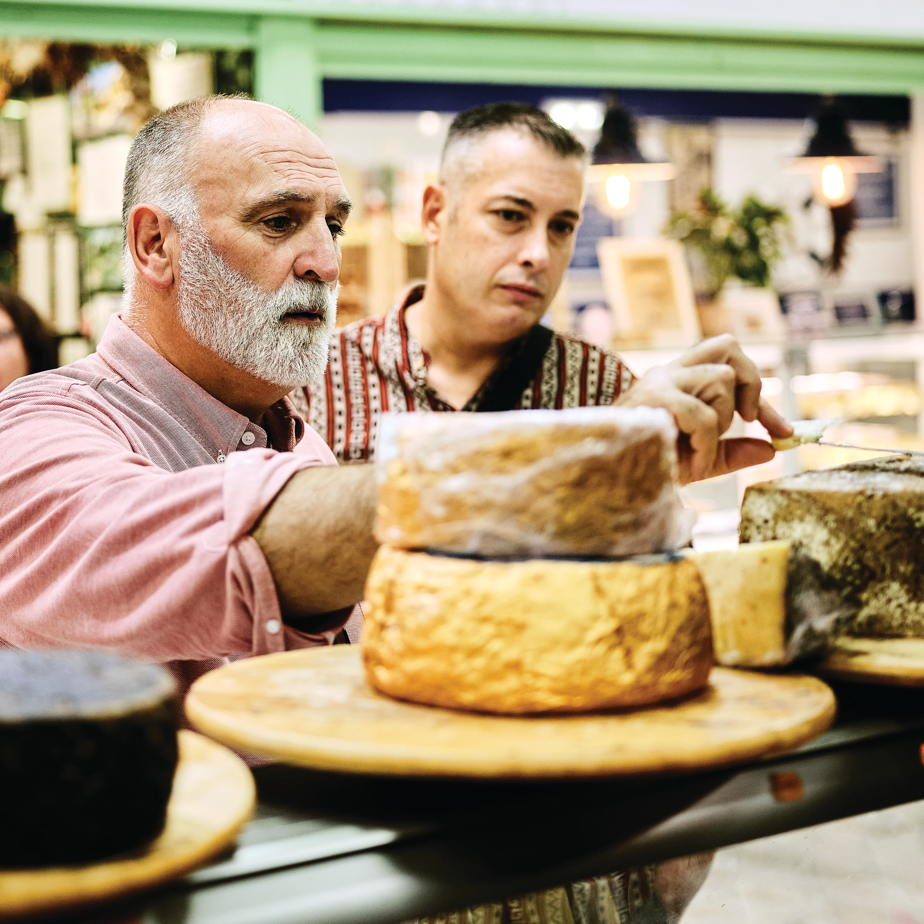 Two men examine large wheels of aged cheese on a wooden board at an indoor market cheese counter.
