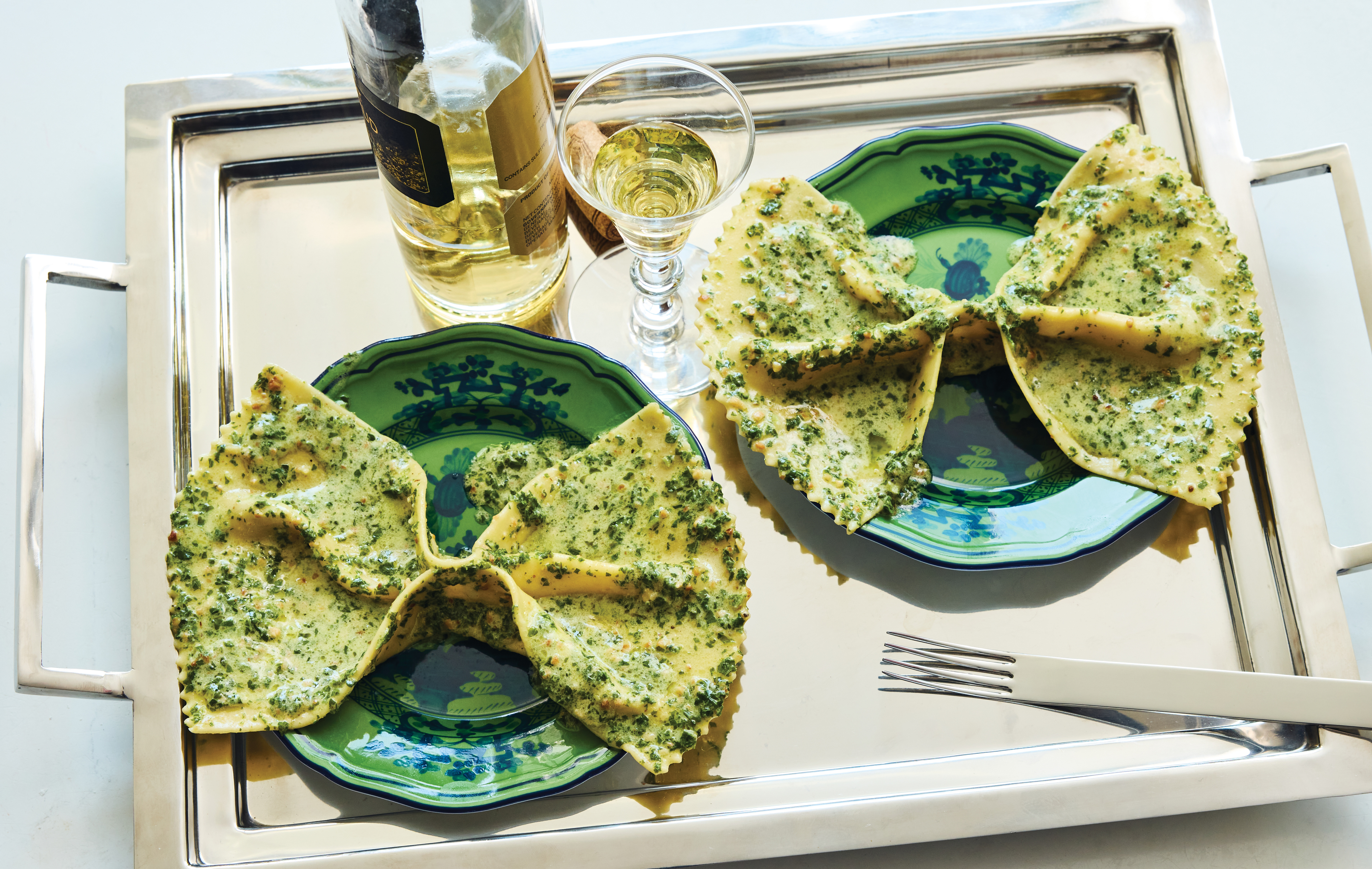 Two plates of pesto-topped ravioli on green patterned plates, served on a silver tray with white wine and a fork.