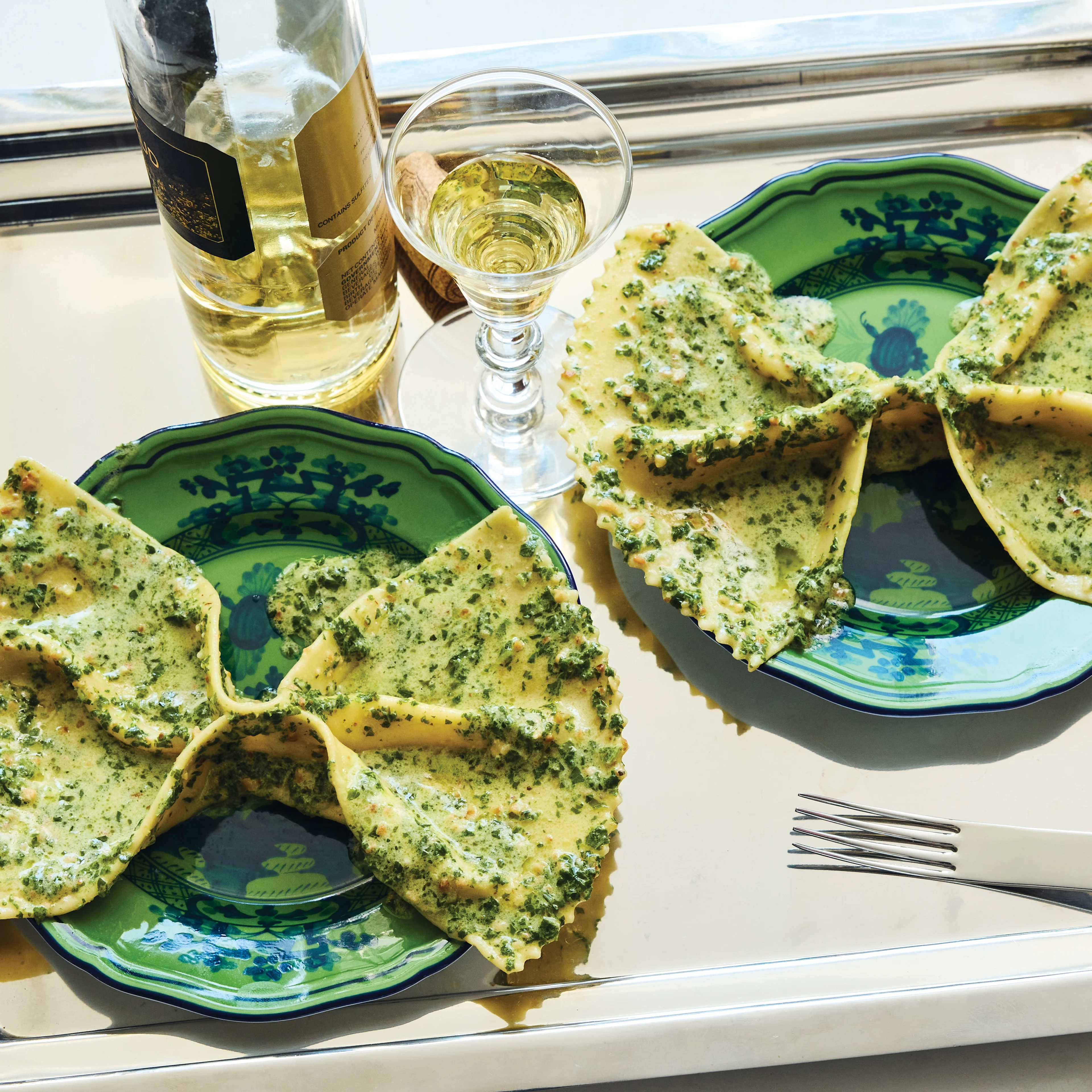 Two plates of pesto-topped ravioli on green patterned plates, served on a silver tray with white wine and a fork.