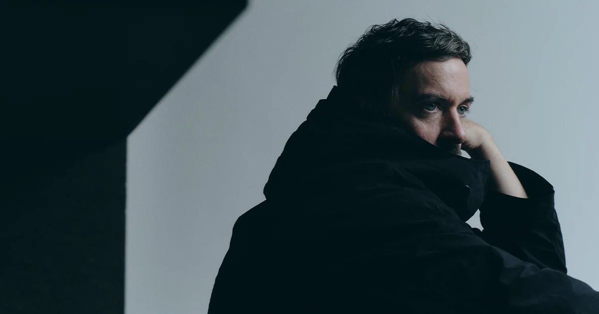 A man in a black jacket glances back over his shoulder against a pale gray background with dramatic shadows.
