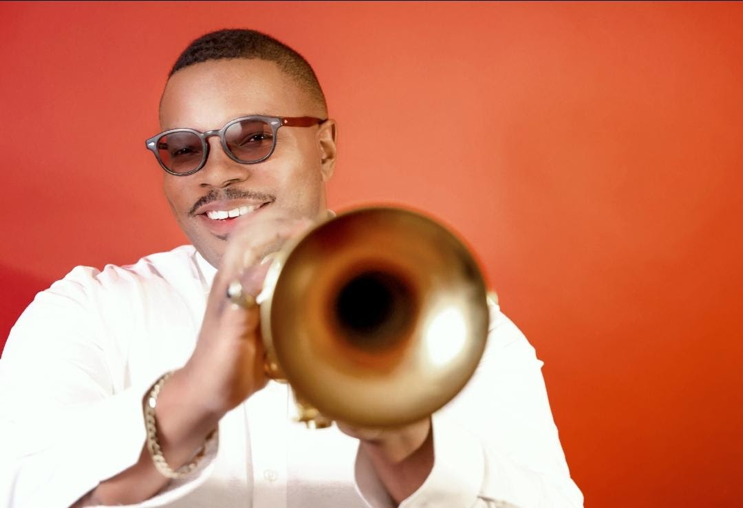 A smiling man in sunglasses and a white shirt holds a gold trumpet toward the camera against a red background.