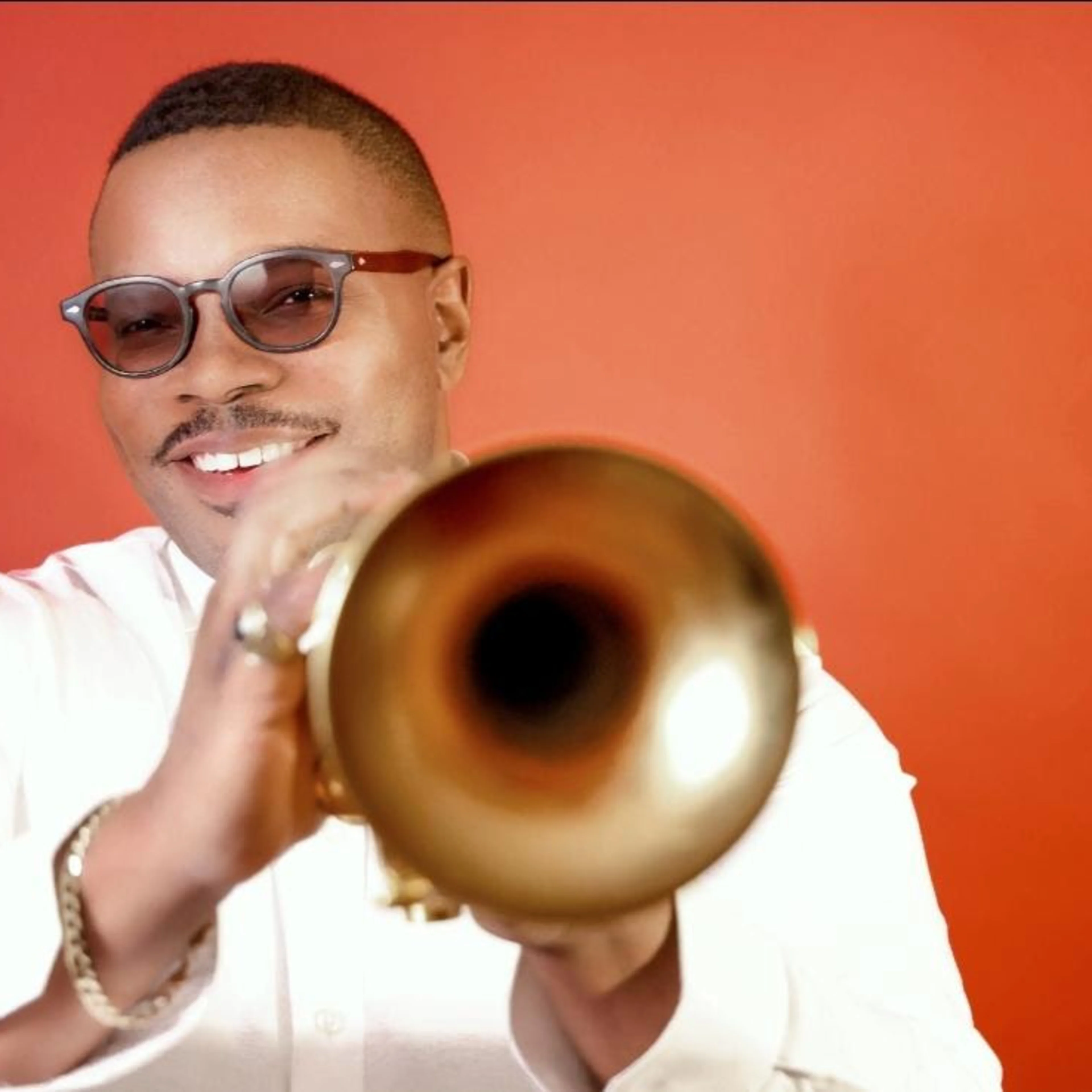 A smiling man in sunglasses and a white shirt holds a gold trumpet toward the camera against a red background.