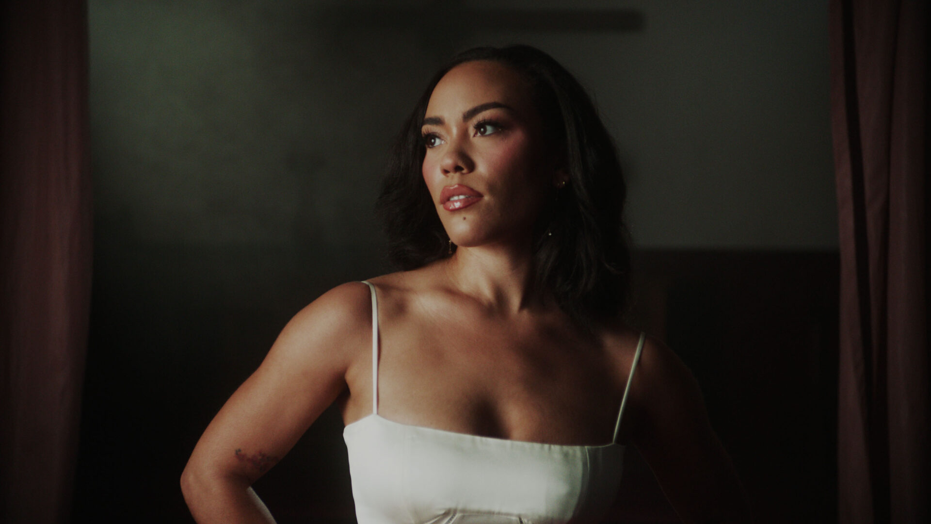 Person with wavy dark hair in white spaghetti strap top, illuminated by soft lighting against a dark background.
