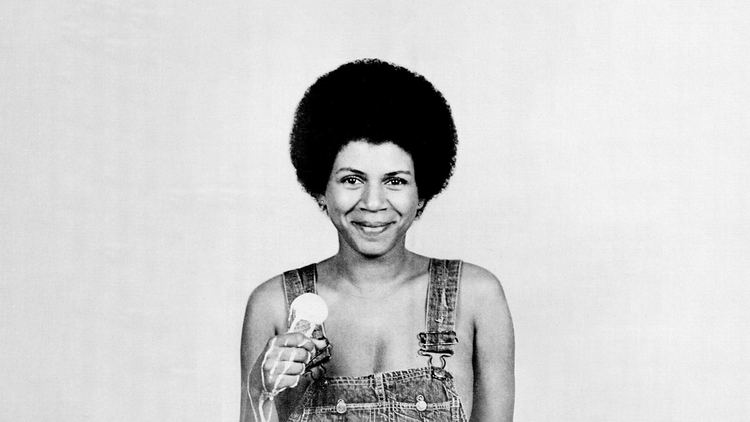 Person with afro hairstyle wearing denim overalls, smiling while holding a microphone in black and white photograph.