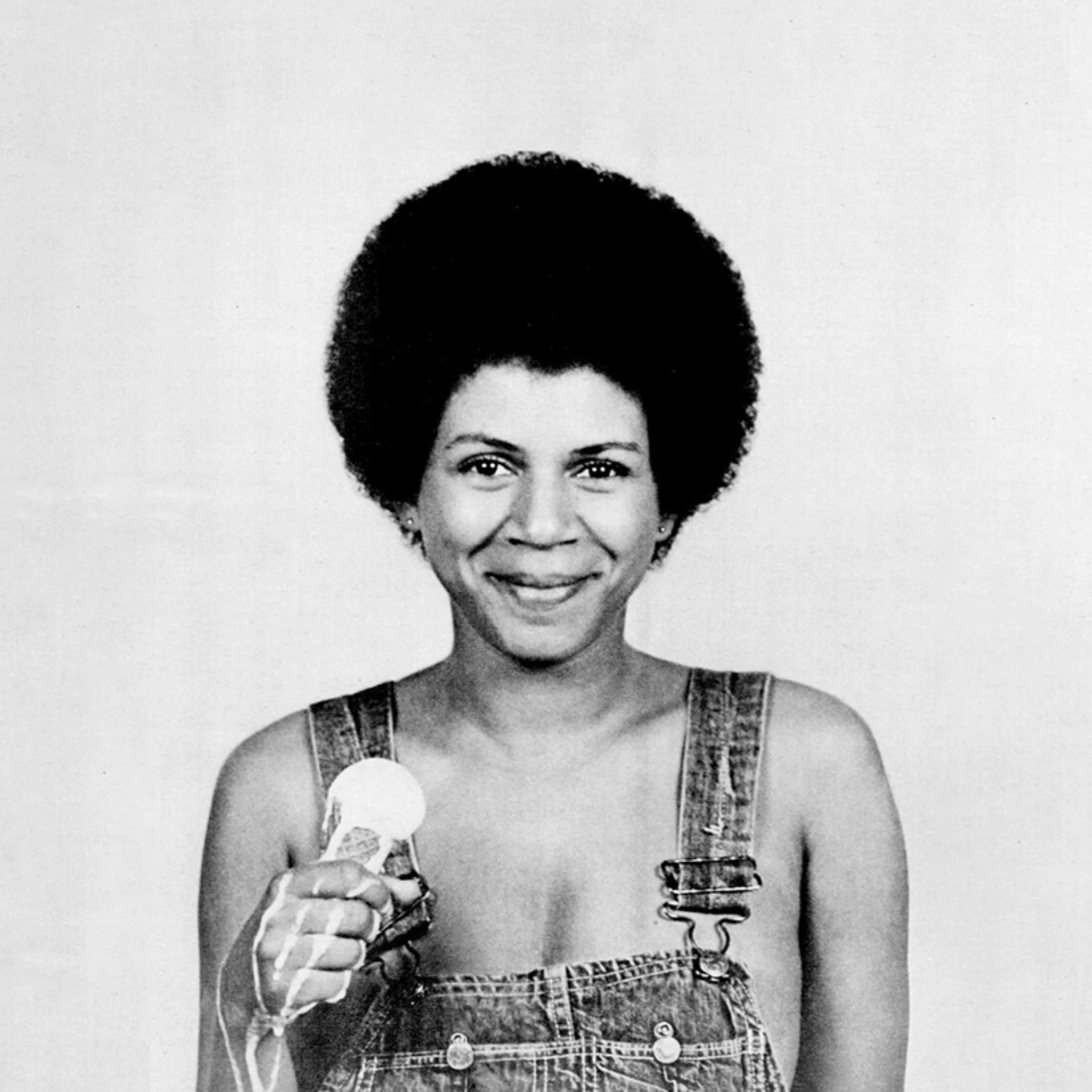 Person with afro hairstyle wearing denim overalls, smiling while holding a microphone in black and white photograph.