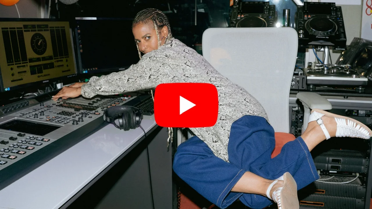 Novena Carmel in radio studio with mixing equipment, YouTube play button overlay, wearing blue pants and patterned top.