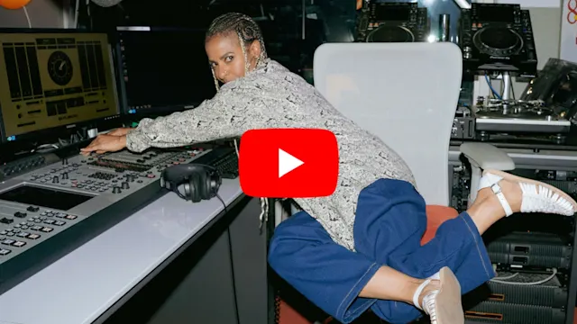 Novena Carmel in radio studio with mixing equipment, YouTube play button overlay, wearing blue pants and patterned top.