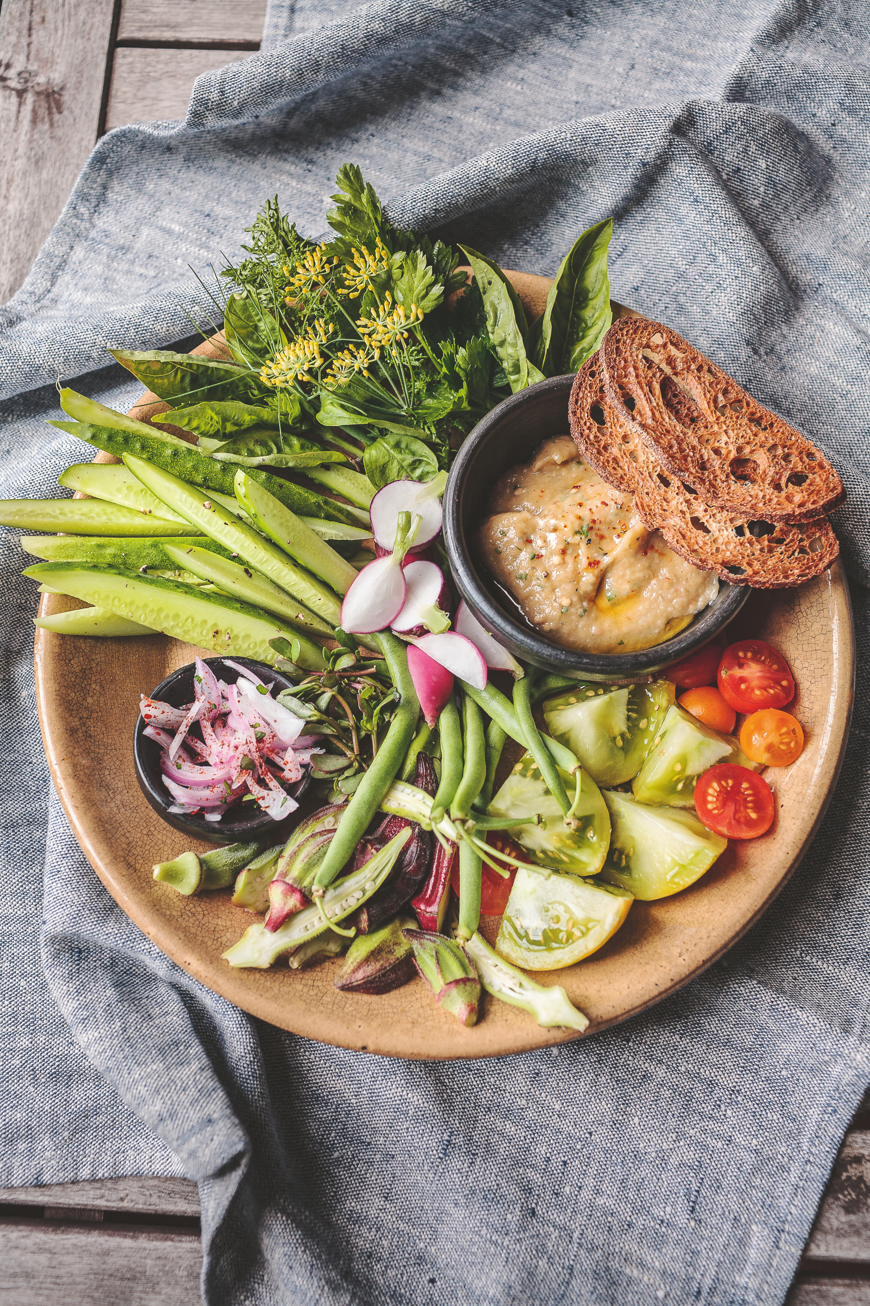 A rustic plate with fresh vegetables, herbs, tomatoes, radishes, green beans, dip, and toasted bread on a gray linen cloth.