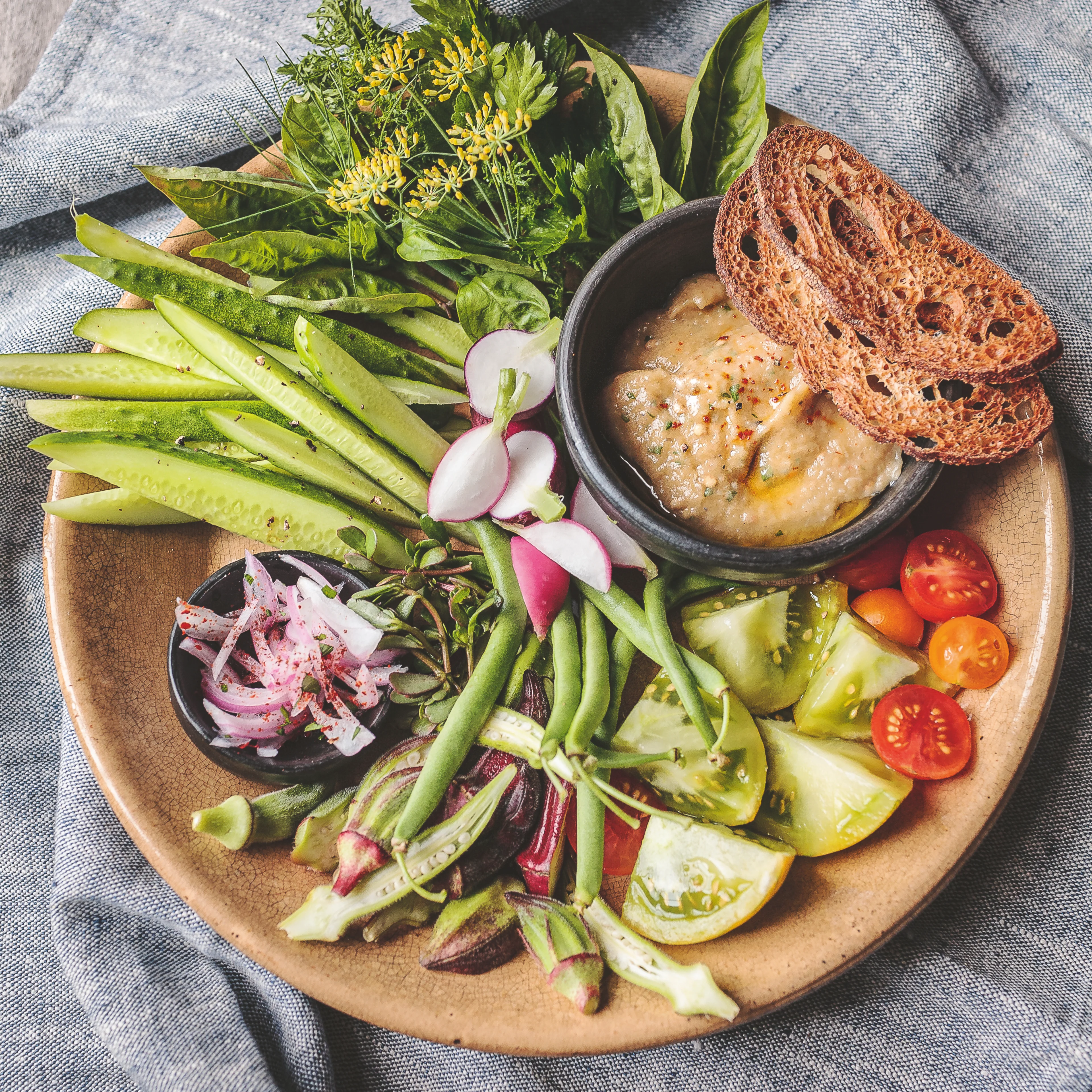 A rustic plate with fresh vegetables, herbs, tomatoes, radishes, green beans, dip, and toasted bread on a gray linen cloth.