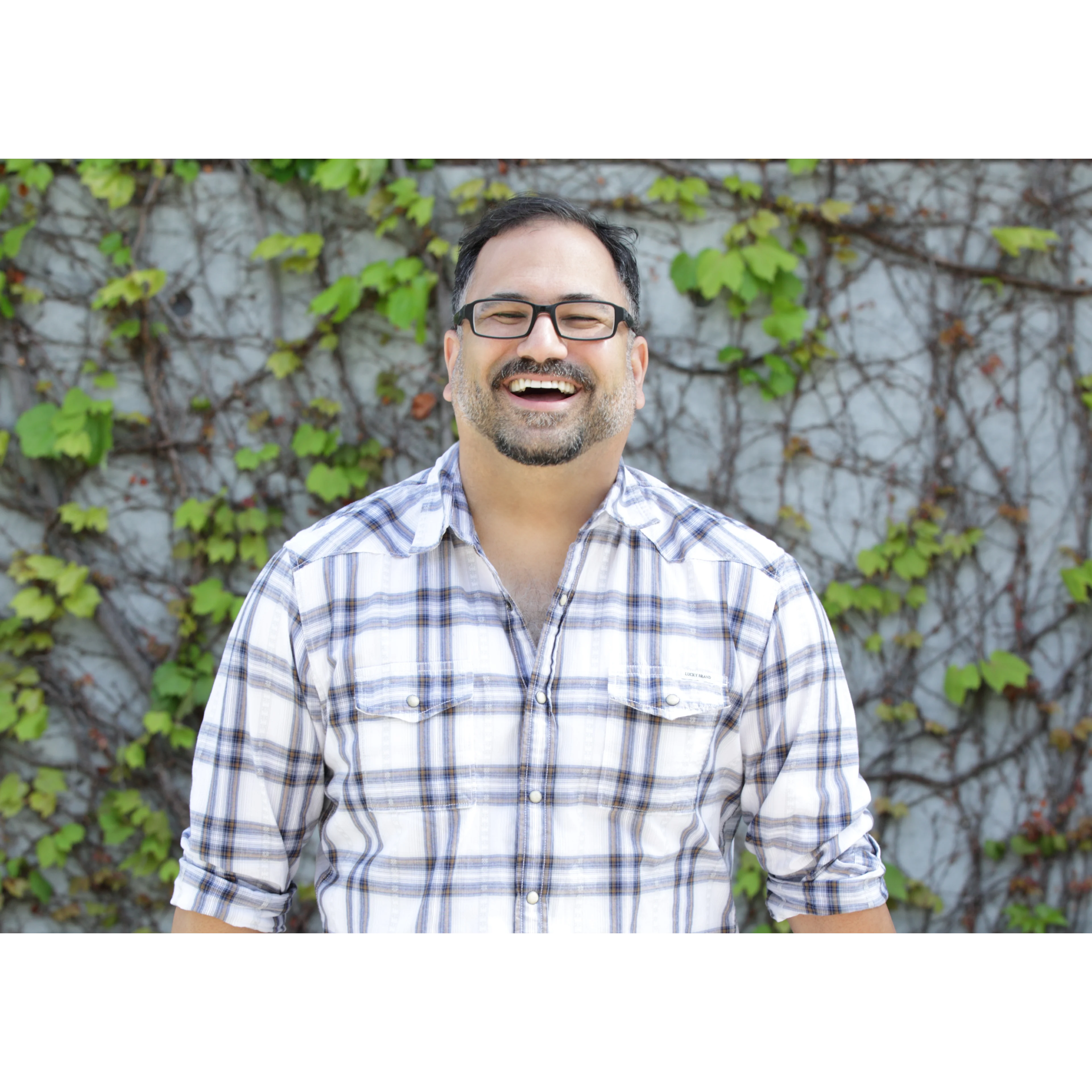 Person smiling broadly wearing glasses and plaid shirt against ivy-covered wall background.