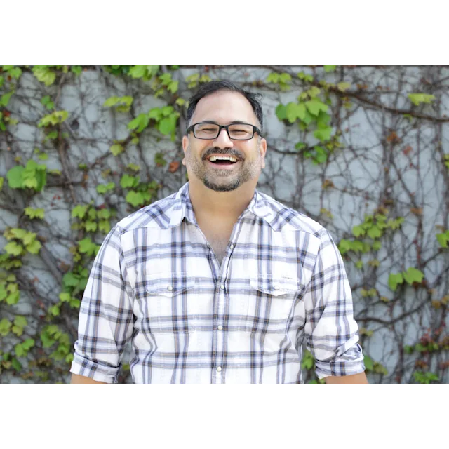 Person smiling broadly wearing glasses and plaid shirt against ivy-covered wall background.