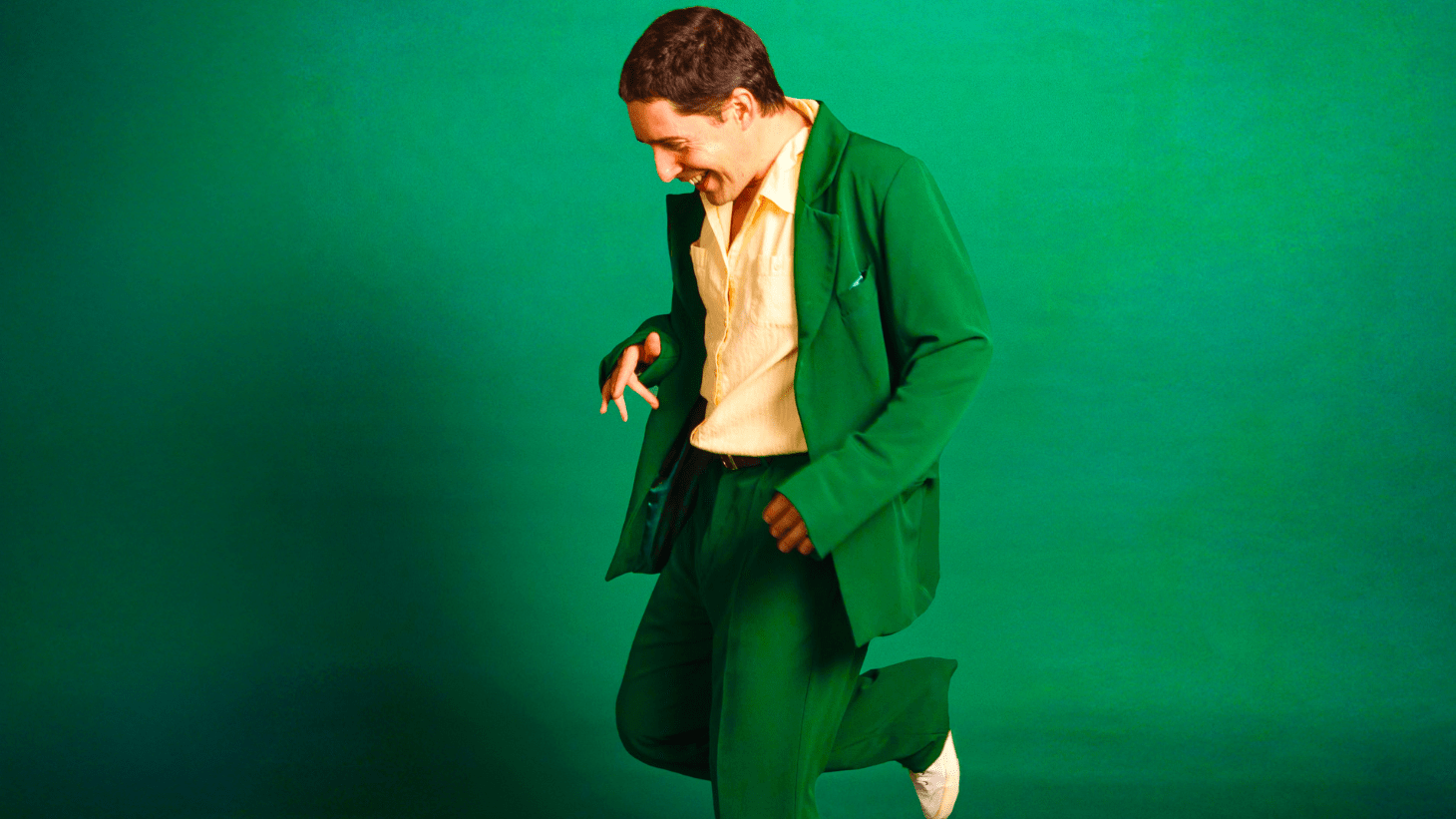 Person in bright green suit with cream shirt smiling and dancing against a vibrant green background.