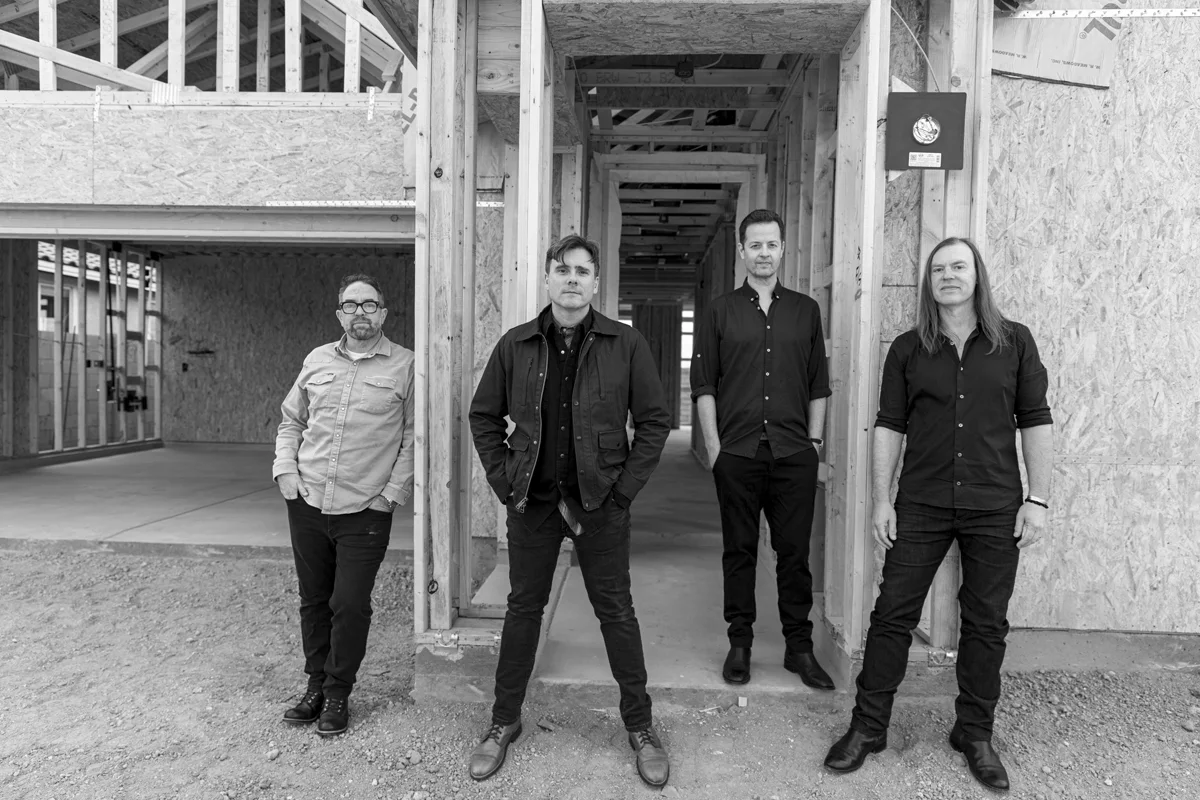 Four men stand in a black and white photo inside an unfinished building with wooden framing and exposed walls.