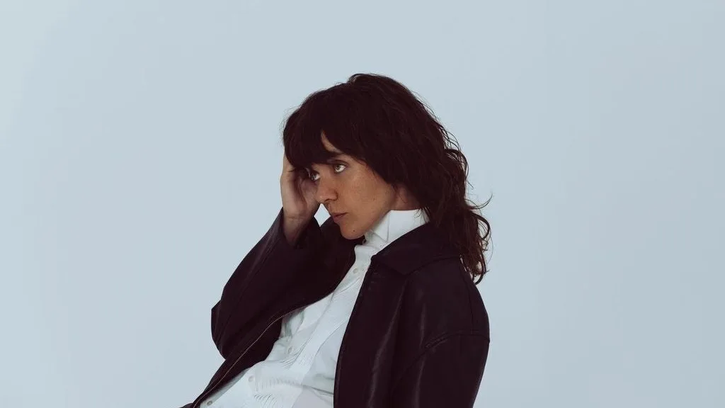 A person with dark shaggy hair rests their hand on their head, wearing a black jacket over a white shirt, against a light blue background.