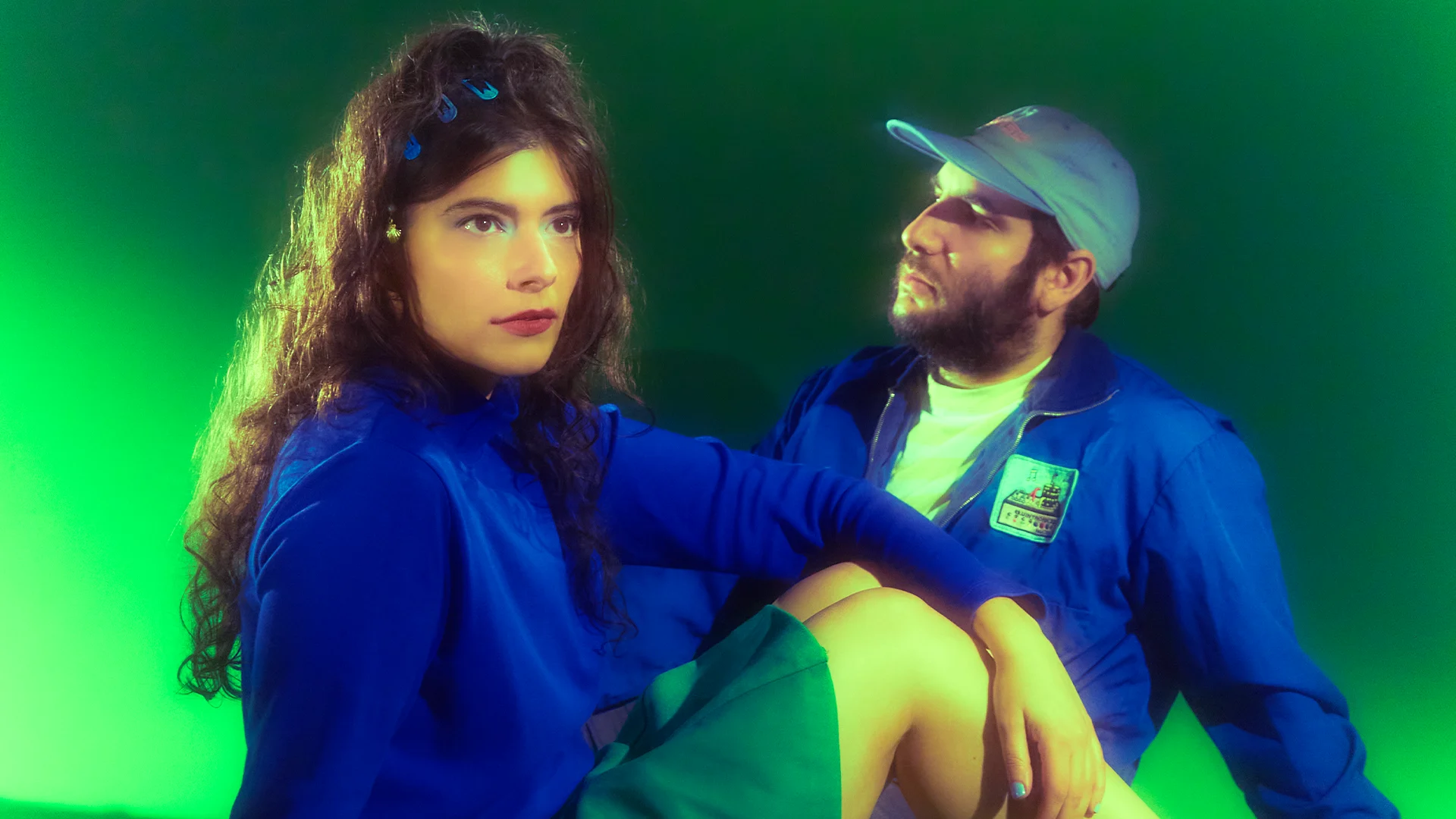 Two people in blue outfits pose against a green background; a woman looks forward while a bearded man in a cap sits behind her.