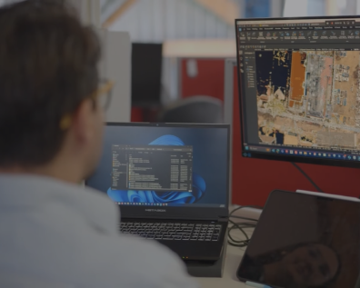 A person working at a desk with two monitors displaying CAD software, showing a detailed site map or aerial drawing.
