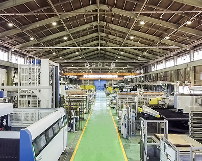 Concept Systems Technologies - Thumbnail Interior of a large industrial manufacturing facility with machinery lining both sides and a high steel-trussed ceiling with overhead lighting.