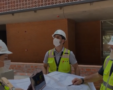 Three construction workers in hard hats and safety vests review blueprints at a building site with brick walls.