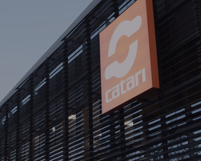Catari - Thumbnail Orange Catari logo sign mounted on a modern building facade with dark vertical slats and interior lighting visible.