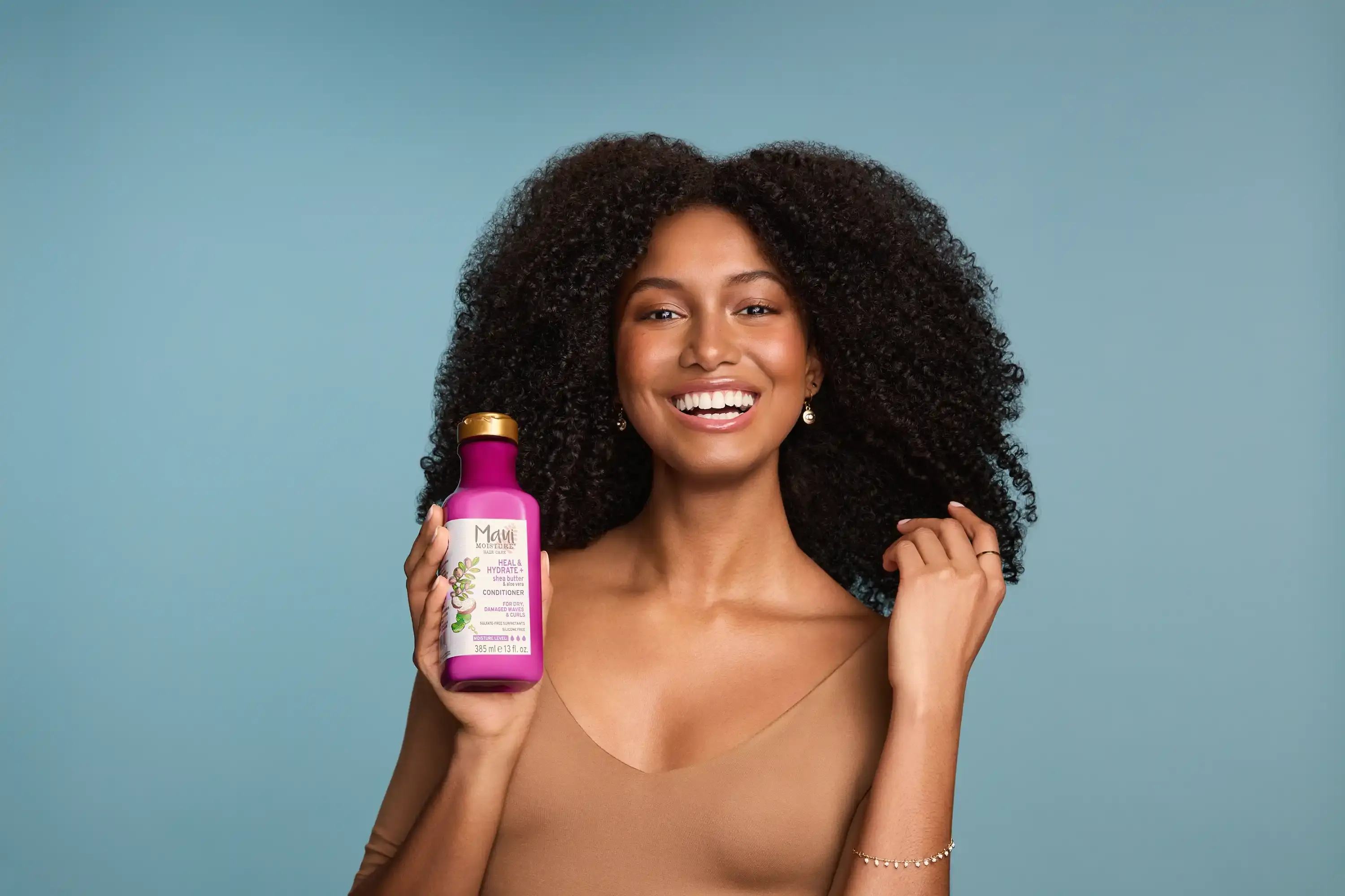 Smiling woman with healthy curls holding Maui Moisture Heal and Hydrate Shea Butter Conditioner