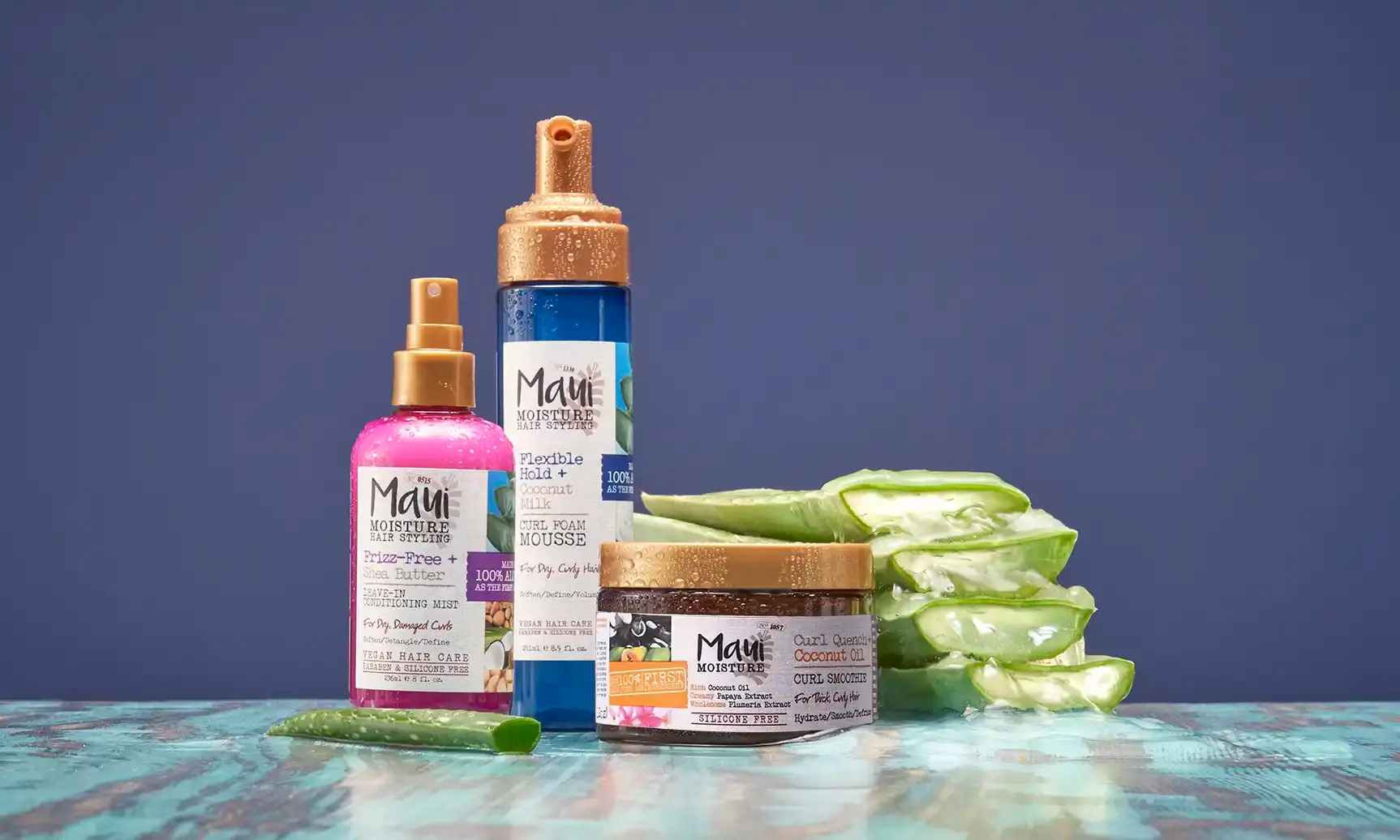  Maui Moisture® hair stylers next to aloe leaves, including conditioning mist, foam mousse, and curl smoothie