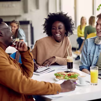 A group of three people laughing at a lunch table enjoying drinks and food