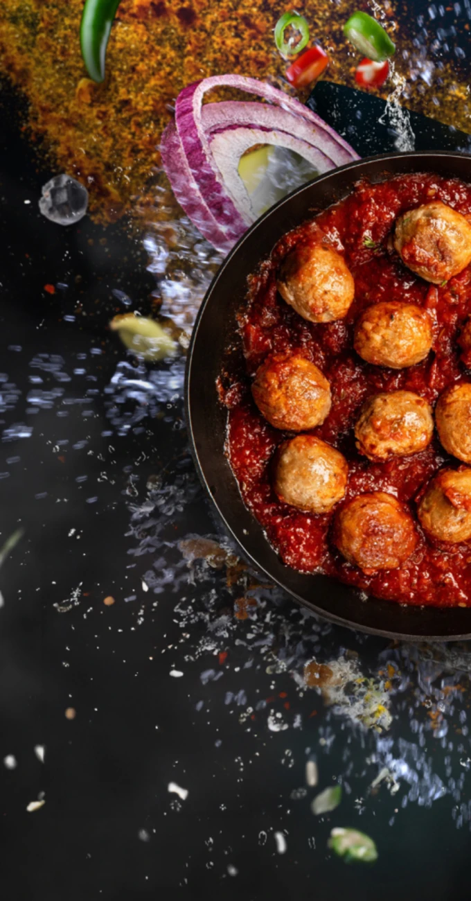 Quorn Swedish Style Meatballs in a saucepan with tomato sauce, with seasoning and vegetables flying into the air in the background
