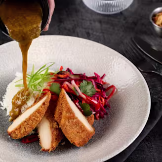 A hand pouring katsu curry sauce on top of chopped up Quorn fillets, rice and salad