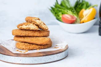 Quorn Vegan Chiqin Strips breaded chicken piled on chopping board, bowl of salad with lettuce, tomato and pepper unfocused in background.