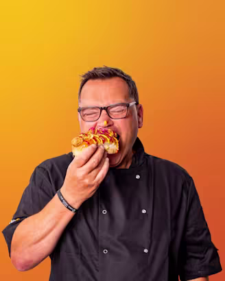 Paul Jennings headshot, Paul wearing glasses while eating a hotdog with mustard and ketchup, dark hair and chef jacket 