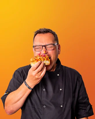 Paul Jennings headshot, Paul wearing glasses while eating a hotdog with mustard and ketchup, dark hair and chef jacket