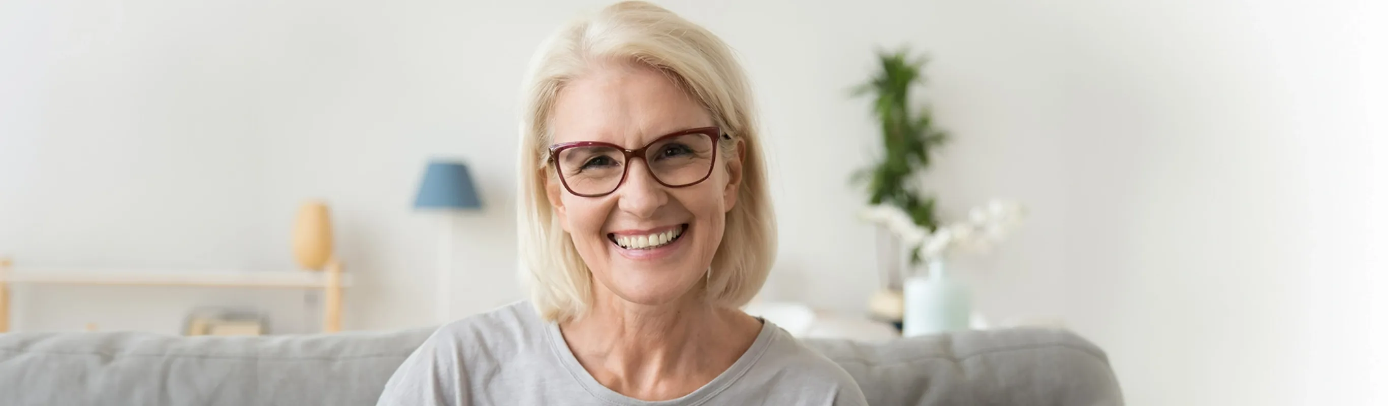 Smiling middle aged mature grey haired woman looking at camera while sitting on sofa in living room