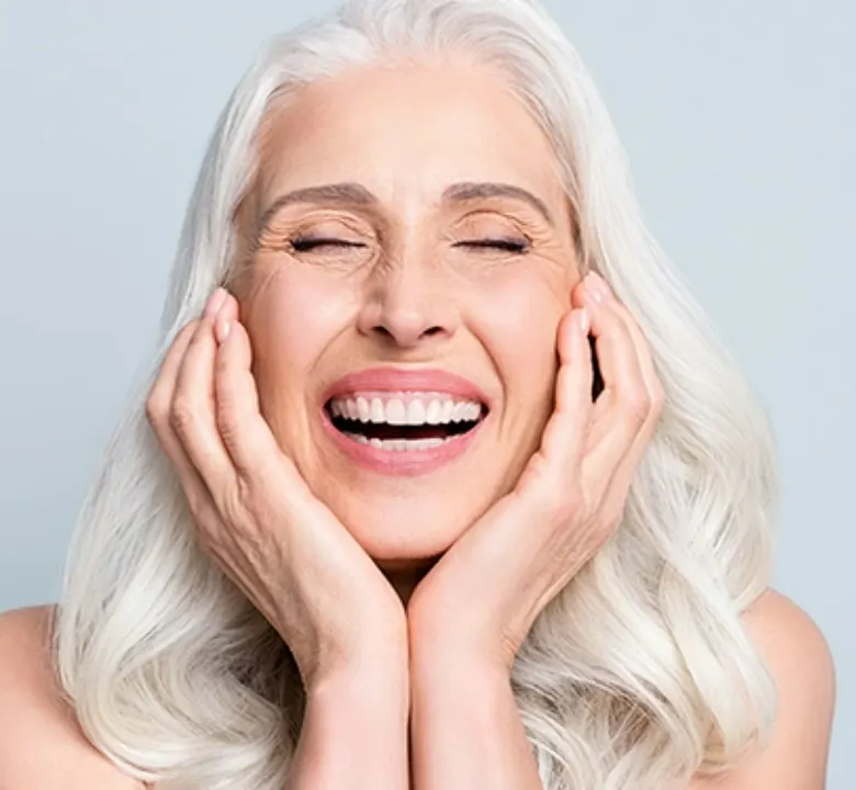 senior woman smiling with grey thick hair and her hands are placed on her cheeks as her eyes are closed in front of a grey background