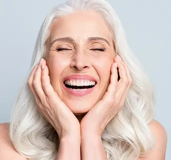 senior woman smiling with grey thick hair and her hands are placed on her cheeks as her eyes are closed in front of a grey background