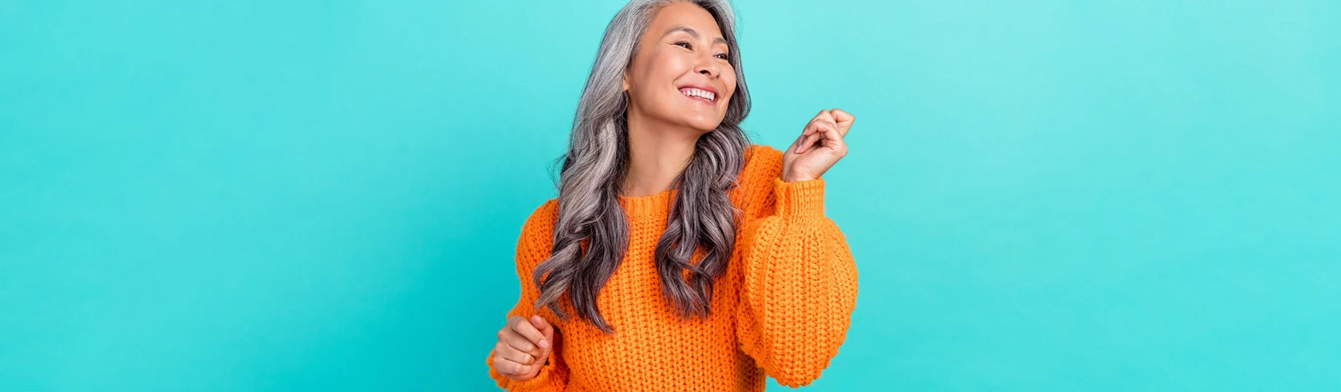 older woman with curly gray hair smiling and dancing wearing an orange sweater in front of a teal background