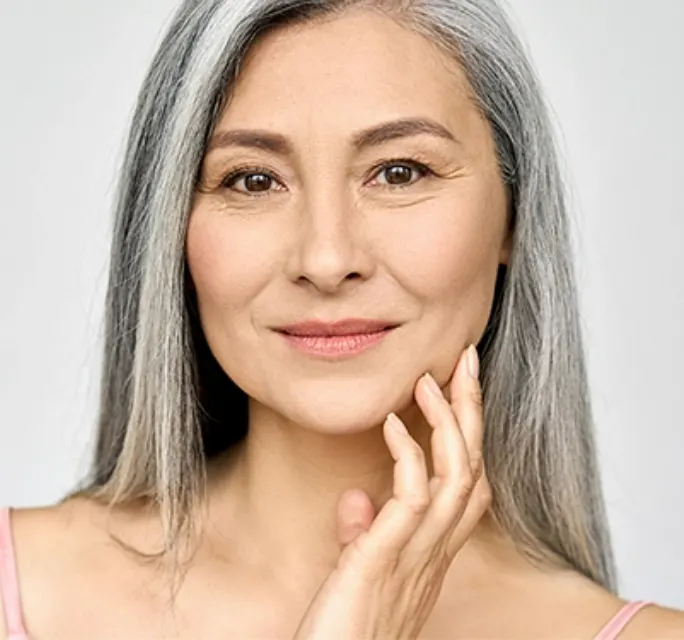 older woman with thin gray hair smirking with her hand grazing her cheek