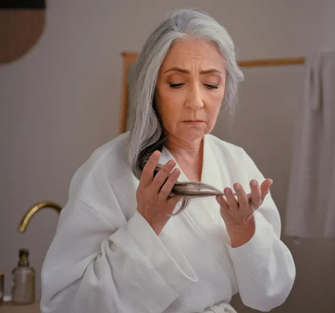 Senior Caucasian woman wearing a bathrobe, gently touching her gray, long, smooth hair, expressing concern about hair loss, thinning hairline, baldness, and the effects of aging, such as alopecia.