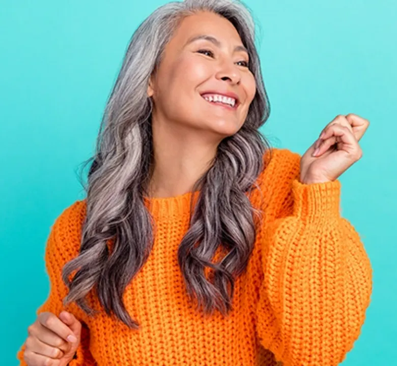 older woman with curly gray hair smiling and dancing wearing an orange sweater in front of a teal background