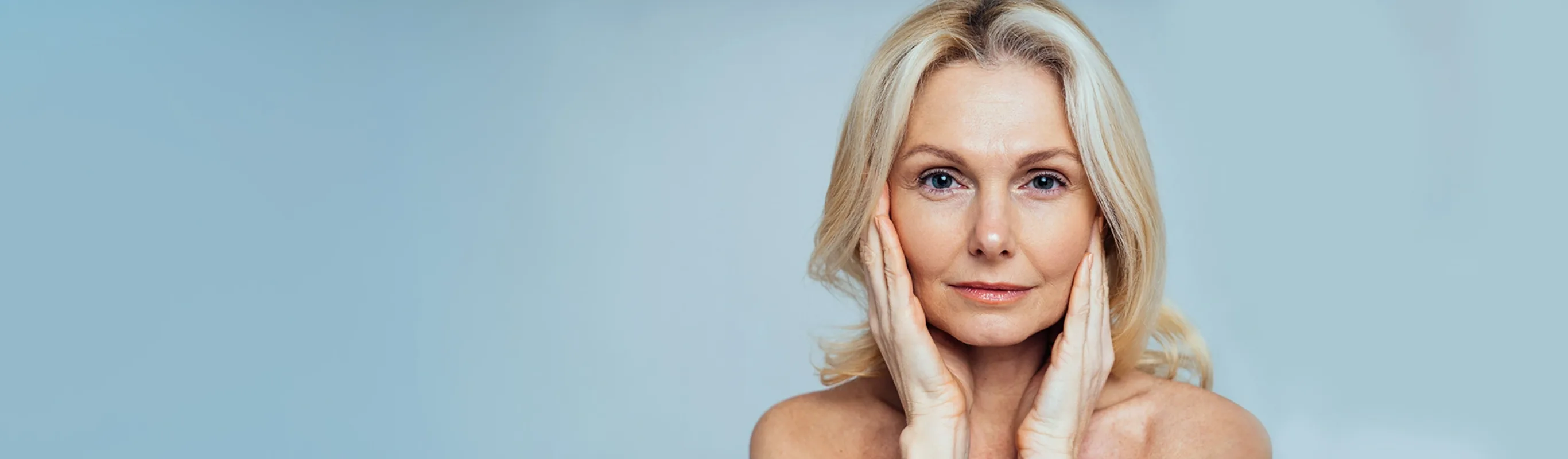 Senior woman with blonde and grey hair her hands on her cheeks posing in front of a blue background