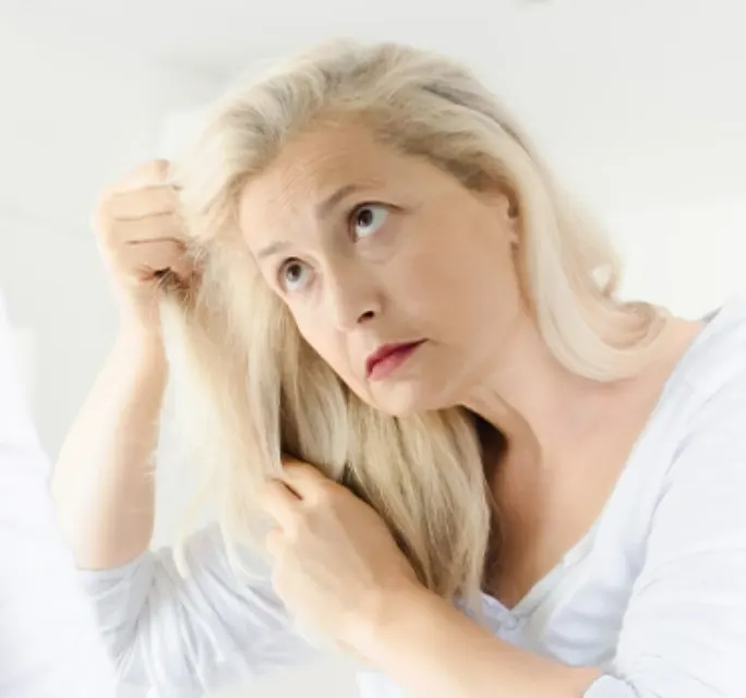 Upset senior woman looking through her thin hair as she stands in her bedroom looking in the mirror